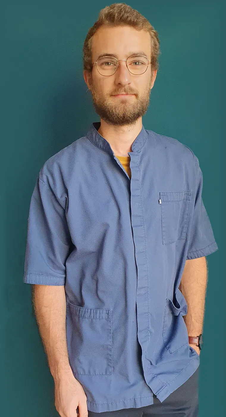 Ronan, en blouse bleue, debout devant le mur bleu-vert de son cabinet